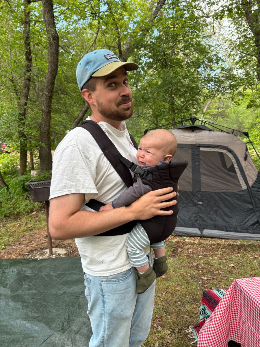 Dave carrying his baby in a chest carrier at a wooded campsite with a tent behind them