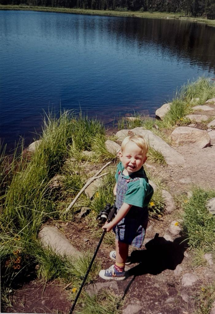 Toddler Dave in overalls gripping a fishing pole beside a mountain lake